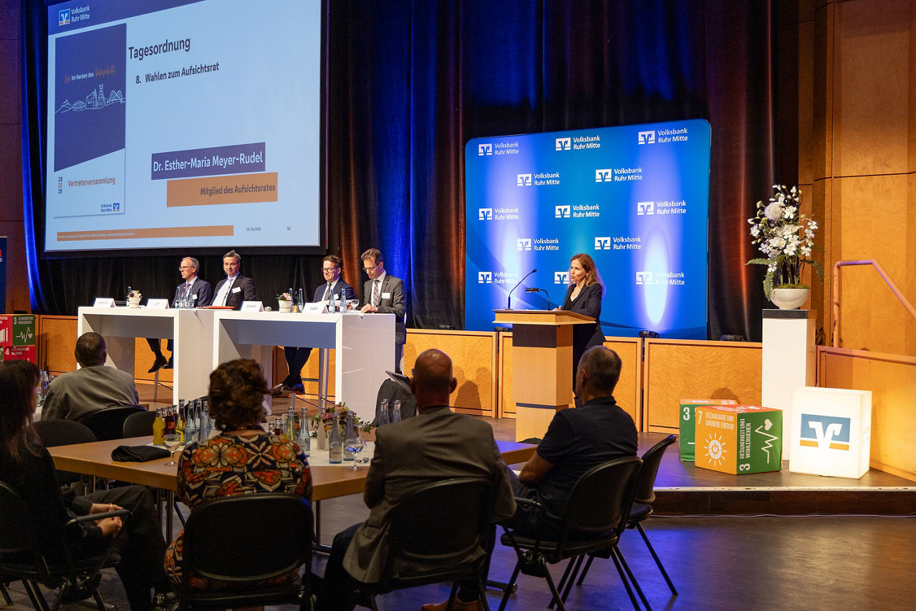 Man sieht das innere der Stadthalle Gladbeck, inklusive der Bühne und der großen Projektorleinwand. Auf der Bühne stehen und sitzen fünf Personen, die Frau davon spricht gerade am Rednerpult. Im Vordergrund sind einige sitzende Zuhörer zu sehen. Es handelt sich um eine Momentaufnahme der Vertreterversammlung der Volksbank Ruhr Mitte.