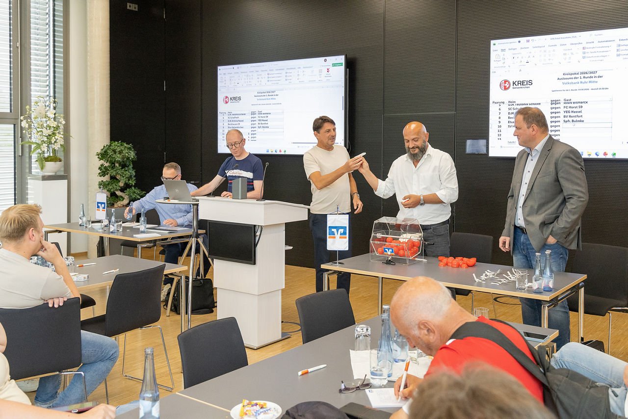 Aufnahmen aus dem Forum der Volksbank Ruhr Mitte im Zuge der Auslosung der Erstundenpartien im Kreispokal Gelsenkirchen. Man sieht einige Zuschauervon hinten in den Raum hineinfotografiert. Vorne stehen vier Männer am Rednerpult und führen die Ziehung durch. Hinten an der Wand hängen zwei Monitore, auf denen die Ergebnisse zu sehen sind.