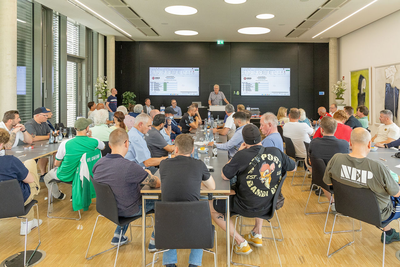 Aufnahmen aus dem Forum der Volksbank Ruhr Mitte im Zuge der Auslosung der Erstundenpartien im Kreispokal Gelsenkirchen. Man sieht die gesamte Zuschauerzahl von hinten in den Raum hineinfotografiert und vorne steht ein Mann am Rednerpult. Hinter dem Mann an der Wand hängen zwei Monitore, auf denen die Ergebnisse zu sehen sind.