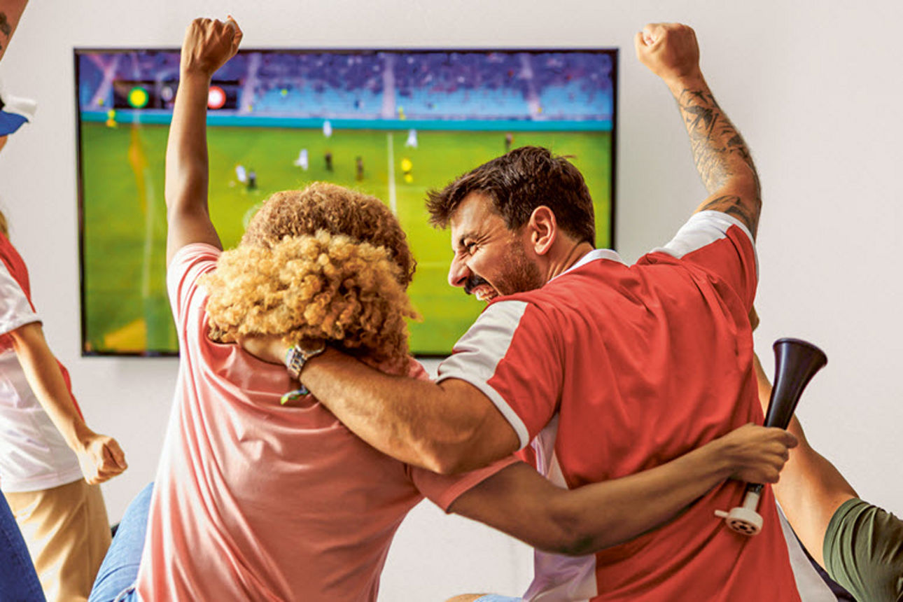 Mehrere Fußballfans jubeln gemeinsam vor einem Fernseher, auf dem ein Spiel läuft, und reißen begeistert die Arme in die Höhe