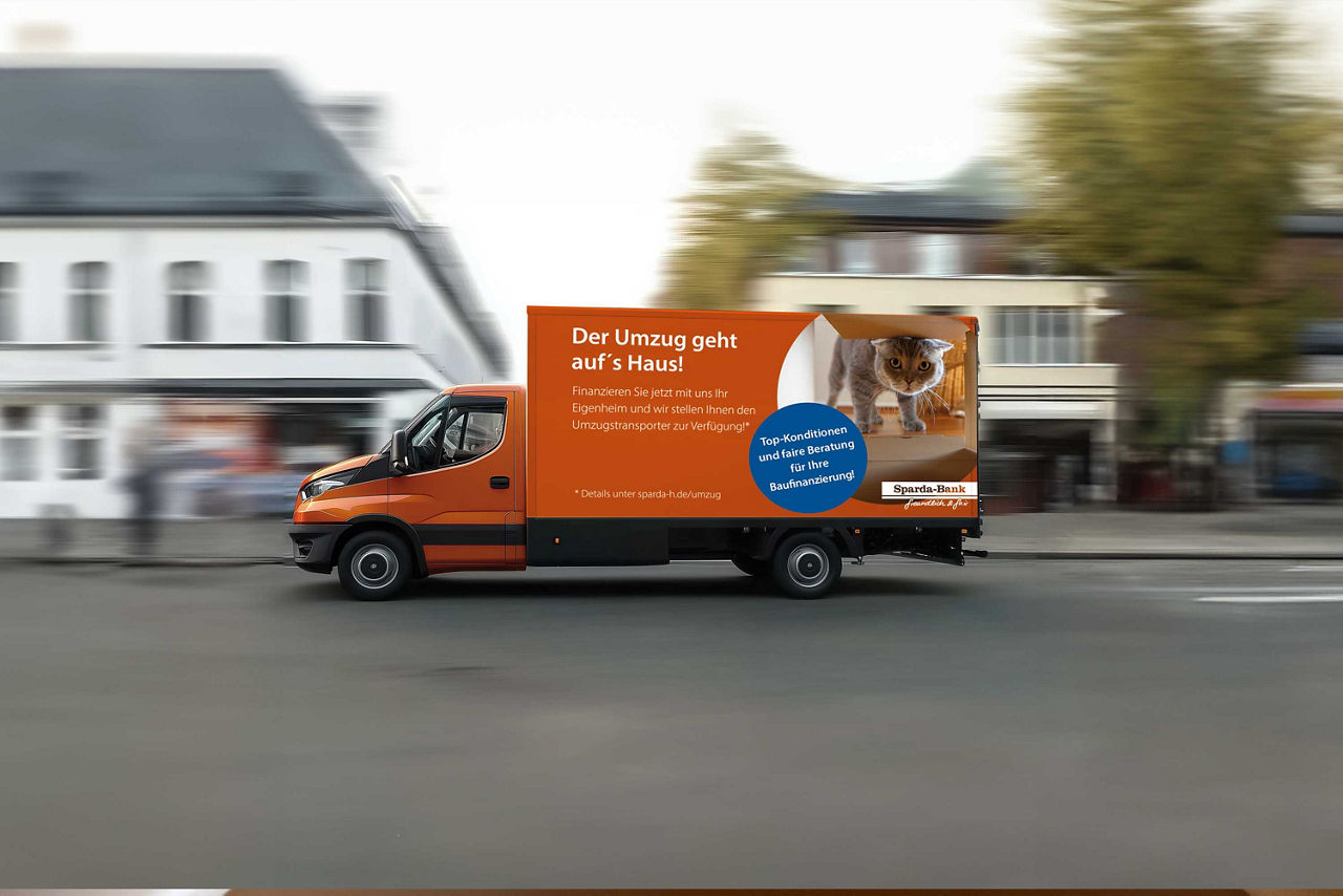 Auf einer Strasse vor zwei Gebäuden fährt ein Transporter mit Werbung der Sparda-Bank Hannover
