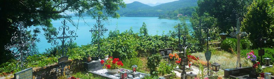 Friedhof mit Blumen und schmiedeeisernen Kreuzen, idyllisch gelegen mit Blick auf einen See und umliegende Berge.