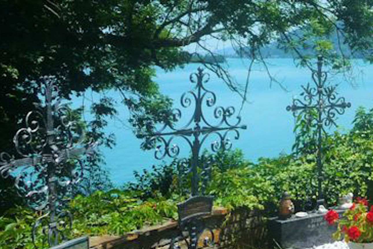 Friedhof mit Blumen und schmiedeeisernen Kreuzen, idyllisch gelegen mit Blick auf einen See und umliegende Berge.