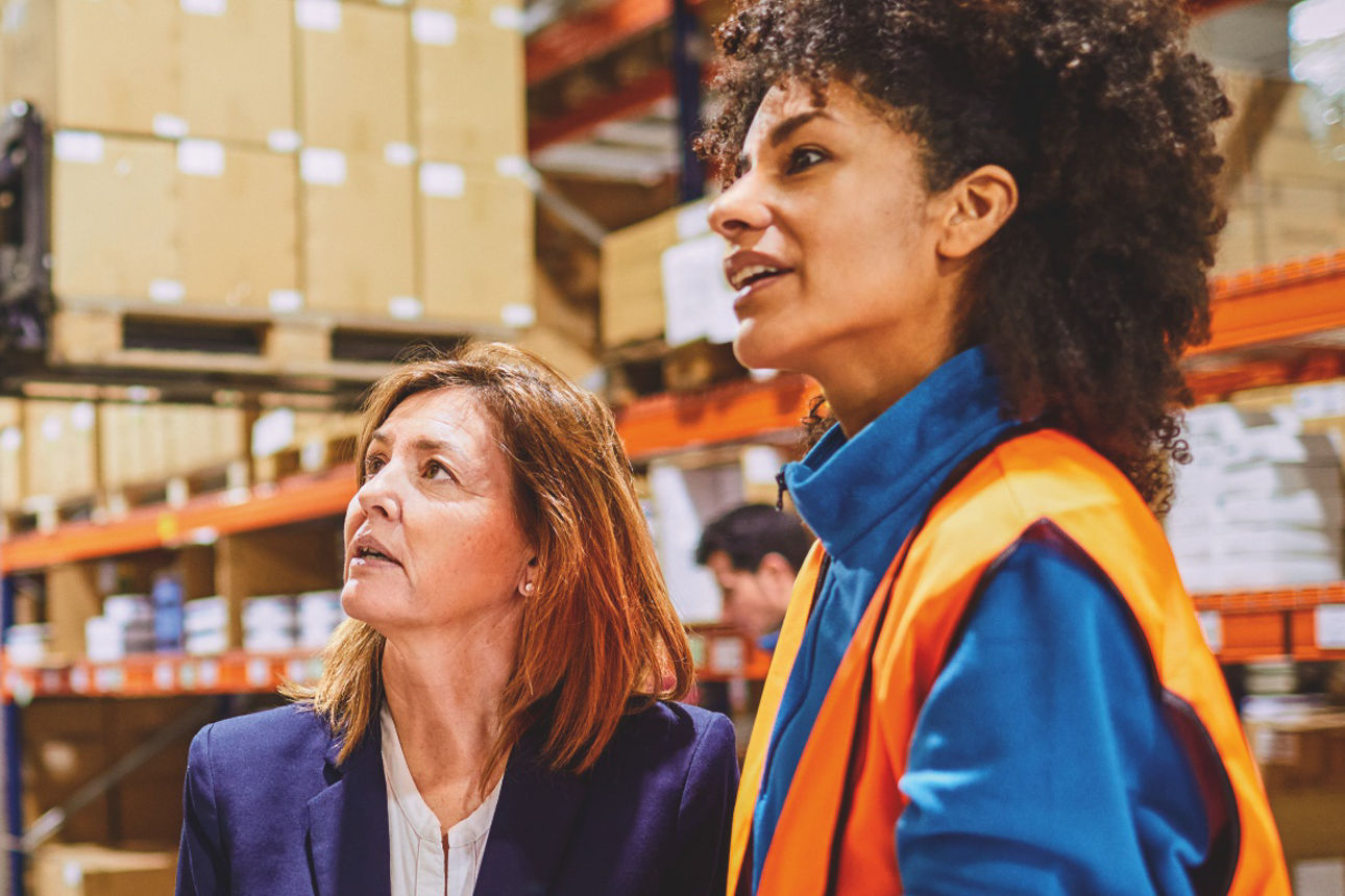 Zwei Personen in Arbeitskleidung mit Warnwesten stehen in einer modernen Produktionsumgebung.