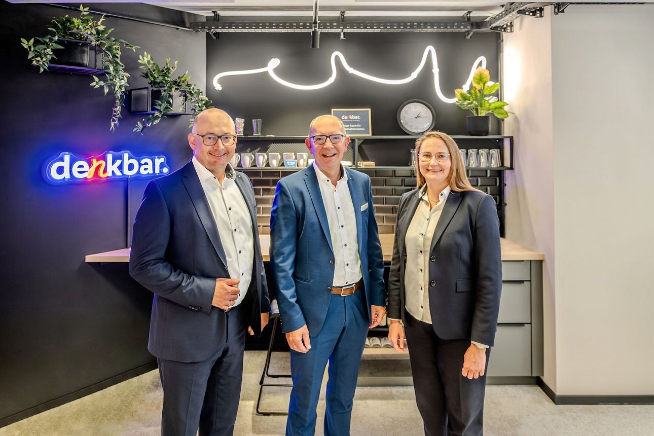 Drei Personen in Business-Kleidung stehen in einer modern gestalteten Kommunikations- und Aufenthaltszone vor einer Küchenzeile. Im Hintergrund sind Pflanzen, Regale, eine Uhr sowie ein beleuchtetes „denkbar.“-Logo und eine dekorative Lichtinstallation zu sehen.