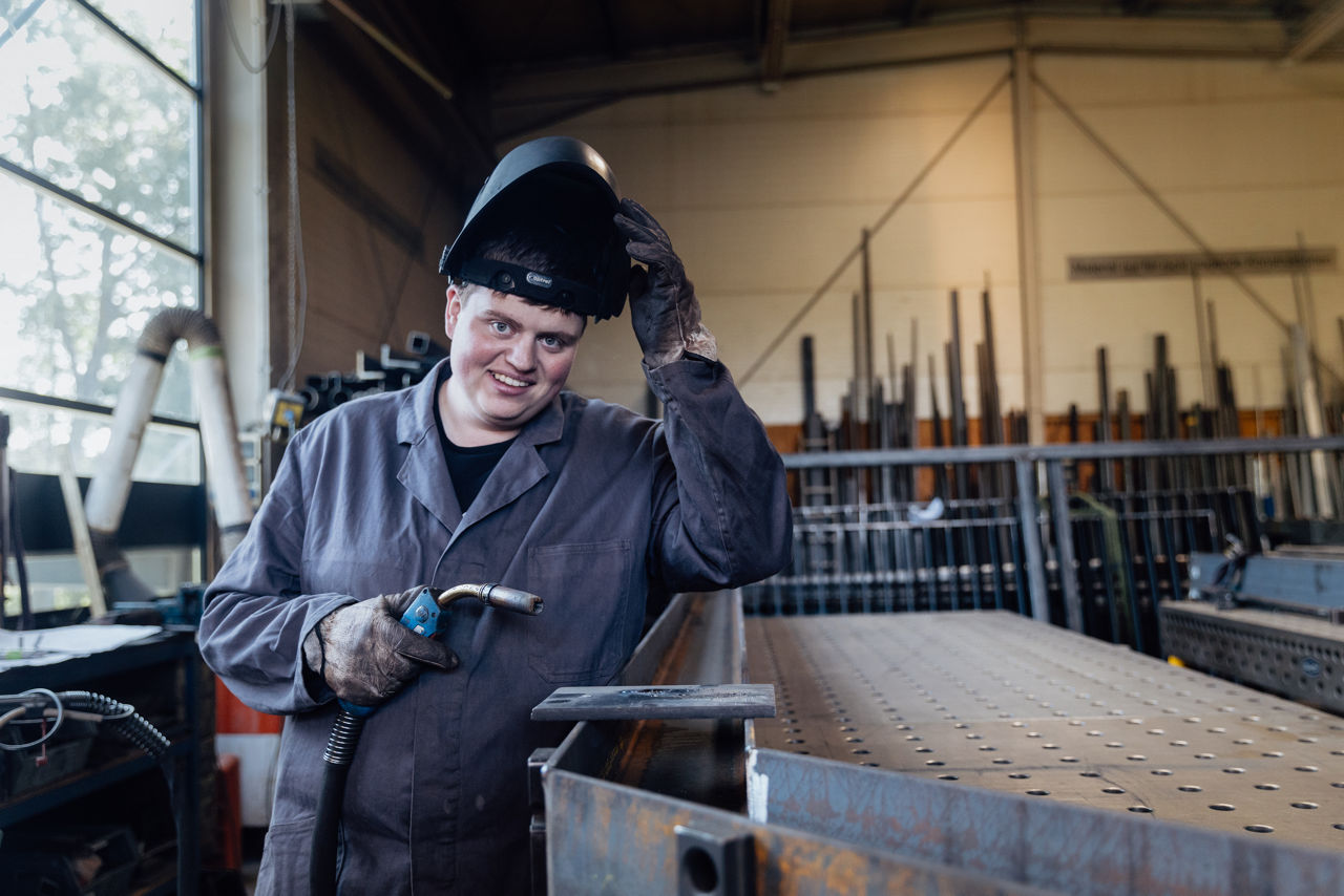 Adam Gilbert steht in der Werkhalle der „AG Metall GmbH“ an einem Metallarbeitstisch mit Schweißgerät und hebt den Schweißhelm an. Die Aufnahme zeigt den Arbeitsbereich während eines Schweißvorgangs im Produktionsumfeld der Metallverarbeitung