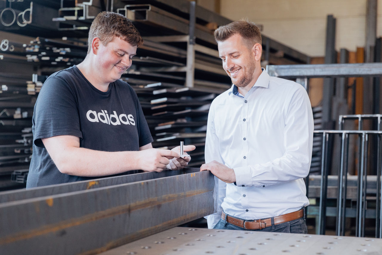 dam Gilbert und Bankberater Timo Barth betrachten in der Werkhalle der „AG Metall GmbH“ ein Metallbauteil und besprechen technische Details. Die Szene zeigt den fachlichen Austausch im Produktionsumfeld mit Fokus auf Metallverarbeitung und Fertigungsqualität