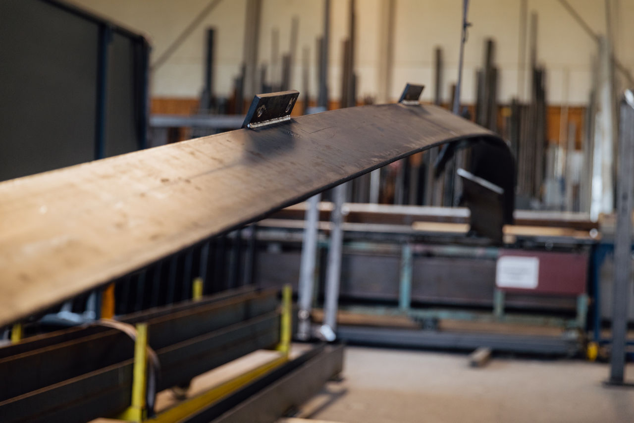 Gebogenes Metallbauteil in der Werkhalle der „AG Metall GmbH“, positioniert vor Maschinen und Lagerelementen. Die Aufnahme zeigt ein bearbeitetes Werkstück für die Weiterverarbeitung im Rahmen der industriellen Metallfertigung