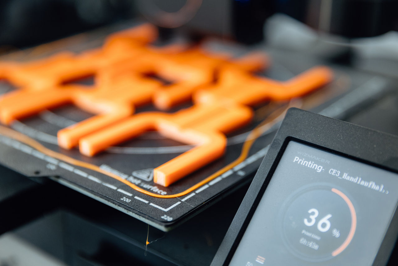 Nahaufnahme einer 3D‑Druckanlage im Produktionsbereich der „AG Metall GmbH“, die orangefarbene Komponenten herstellt. Die Aufnahme zeigt den laufenden additiven Fertigungsprozess und verdeutlicht den Einsatz moderner 3D‑Drucktechnologie innerhalb der industriellen Metall‑ und Komponentenfertigung