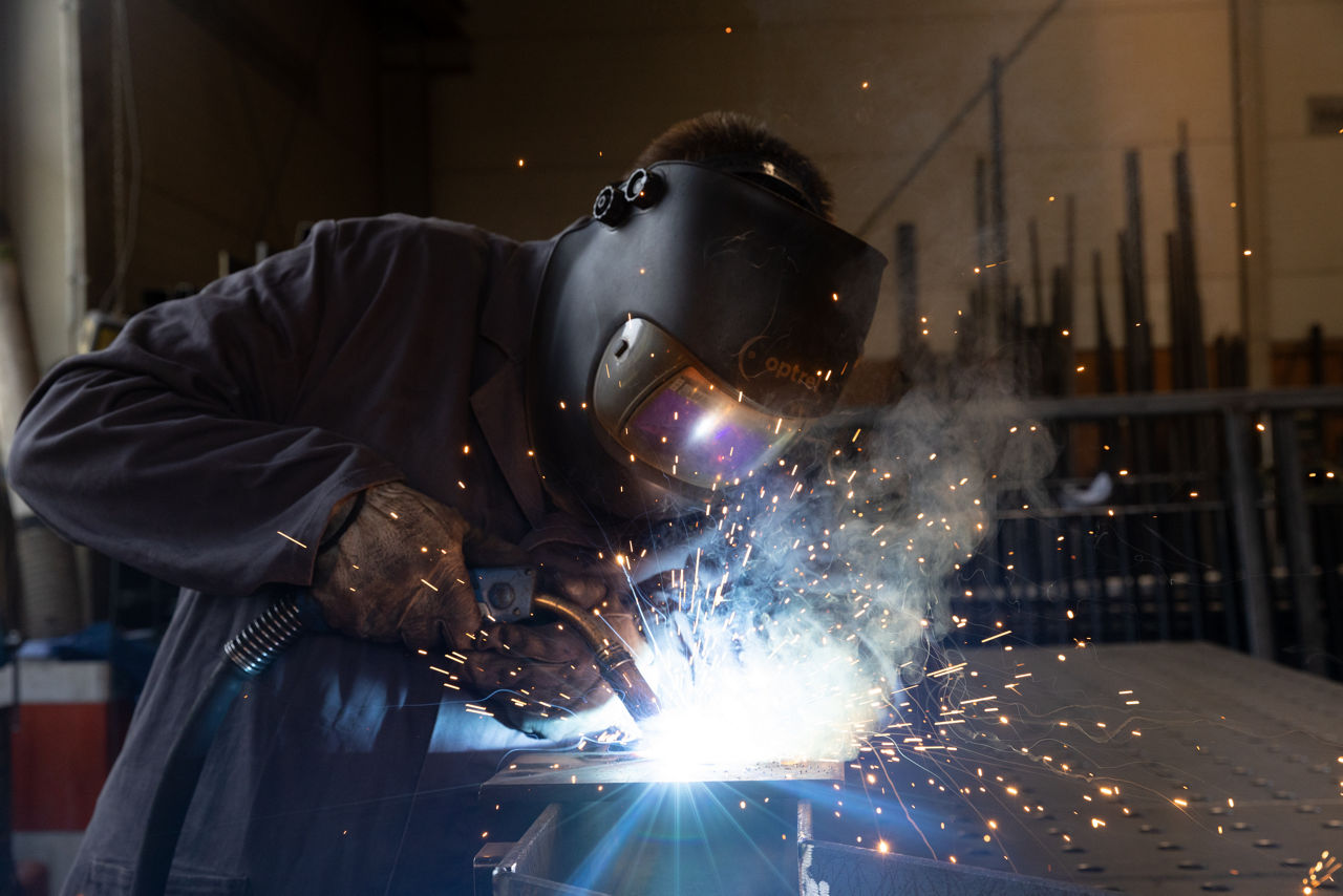 Eine Person führt Schweißarbeiten an einem Metallbauteil in der Werkhalle der „AG Metall GmbH“ aus. Umgeben von Funken, Arbeitsgerät und Metallkonstruktionen zeigt die Aufnahme einen laufenden Schweißprozess im Rahmen der industriellen Metallverarbeitung