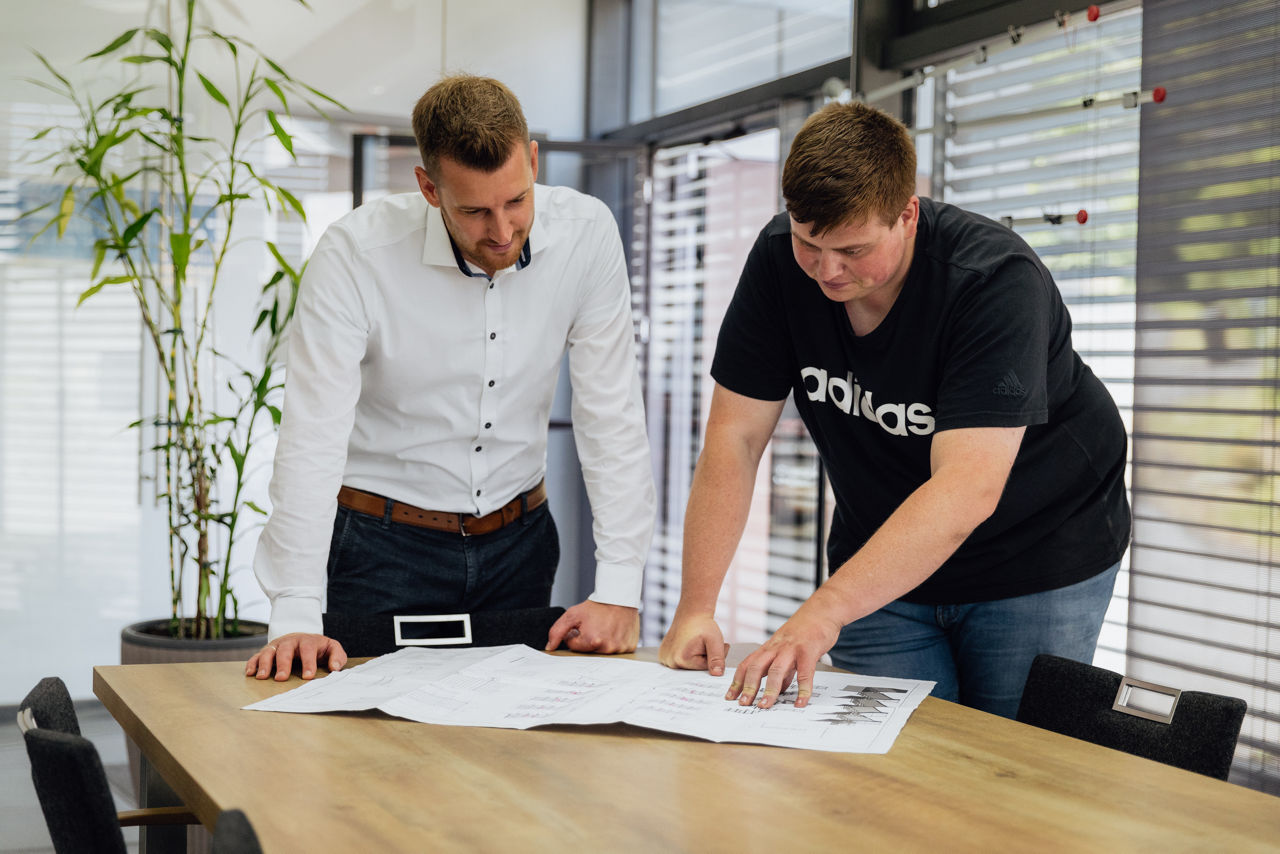 Adam Gilbert und Bankberater Timo Barth besprechen technische Zeichnungen an einem Konferenztisch im Besprechungsraum der „AG Metall GmbH“. Die Aufnahme zeigt den Abstimmungsprozess im Rahmen der technischen Planung und Projektvorbereitung im Unternehmensumfeld