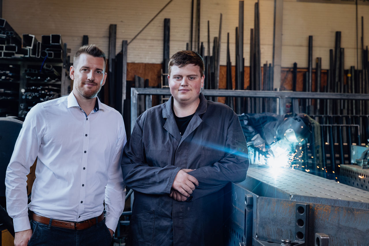 Bankberater Timo Barth und Adam Gilbert stehen in der Werkhalle der „AG Metall GmbH“ vor Metallkonstruktionen. Im Hintergrund finden Schweißarbeiten statt, die den laufenden Fertigungs‑ und Montageprozess im Metallverarbeitungsbetrieb zeigen