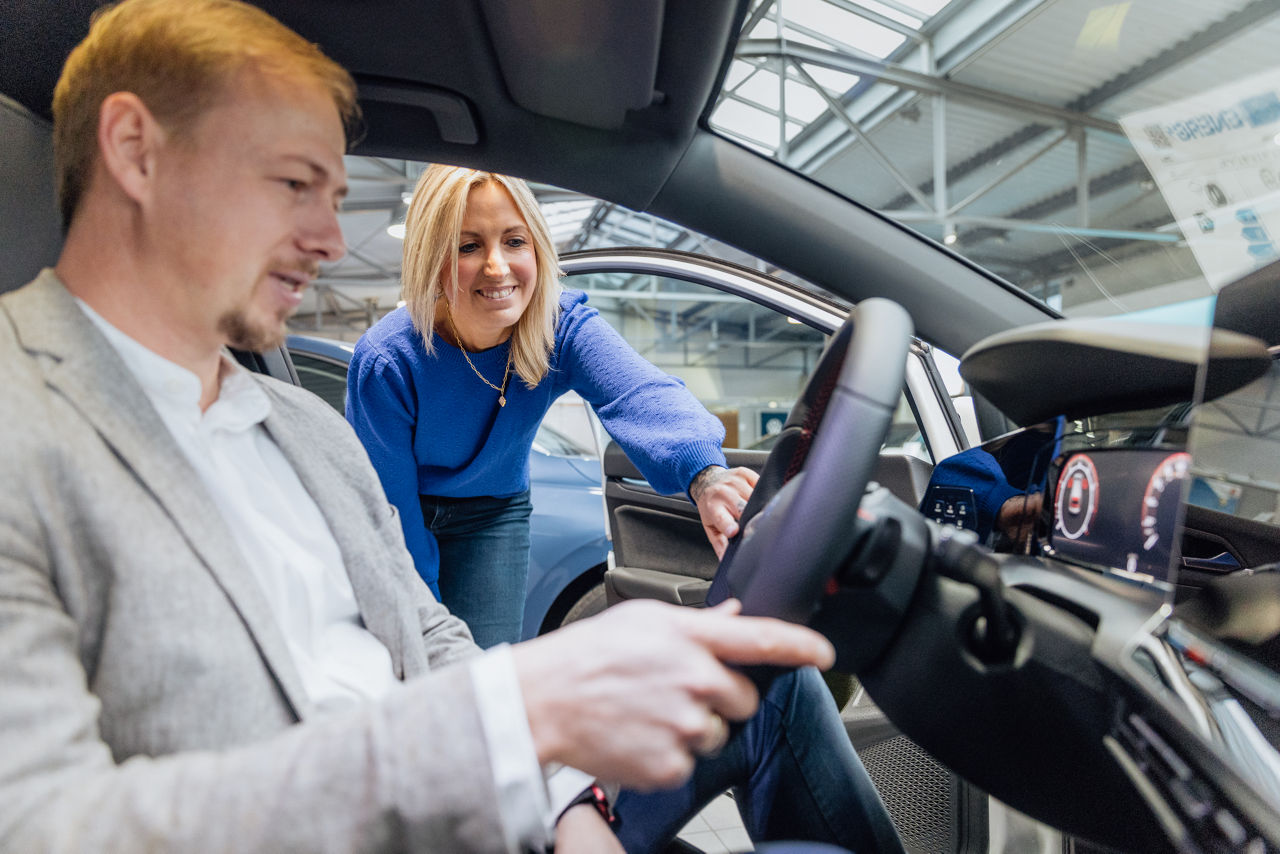 Berater Florian Abschütz und Lisa Woytinnek stellen im Showroom des „Autohaus Kehm“ den Innenraum eines Fahrzeugs vor. Währenddessen werden Multimedia‑ und Fahrzeugfunktionen über die Bedienelemente demonstriert, eingebettet in den Verkaufs‑ und Ausstellungsbereich des Autohauses