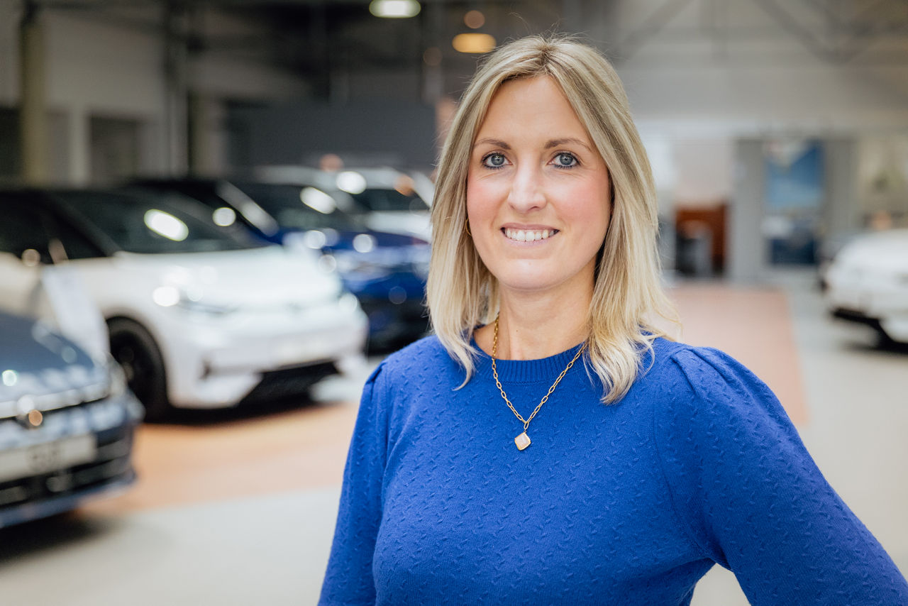 Lisa Woytinnek befindet sich im Innenbereich des „Autohaus Kehm“, umgeben von ausgestellten Fahrzeugen aus dem Volkswagen‑Modellprogramm. Die Aufnahme zeigt den modernen Showroom mit Fahrzeugpräsentation und offen gestalteter Ausstellungsfläche