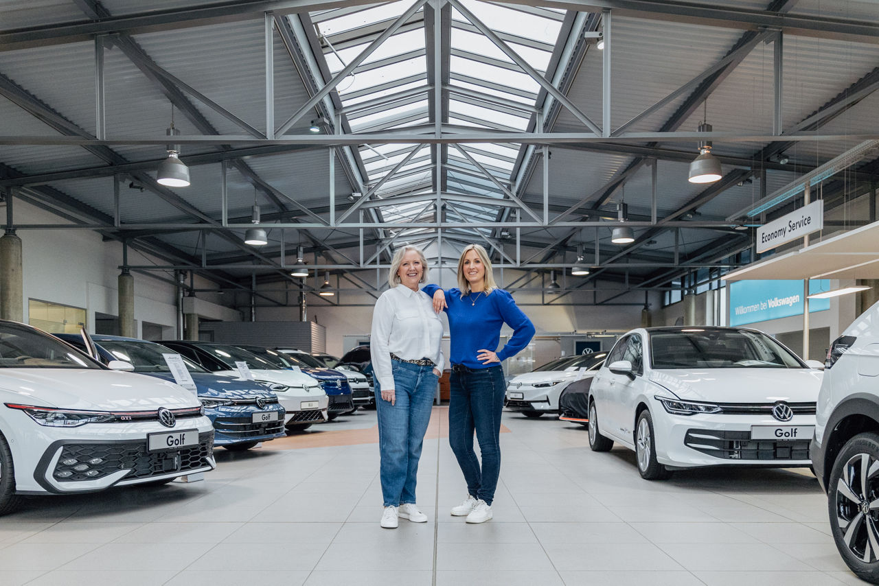 Ulrike Kehm und Lisa Woytinnek befinden sich im Showroom des „Autohaus Kehm“, umgeben von mehreren ausgestellten Fahrzeugen aus dem Volkswagen‑Modellprogramm. Sichtbar sind zudem die offene Decken‑ und Lichtkonstruktion sowie die strukturierte Ausstellungsfläche des Autohauses