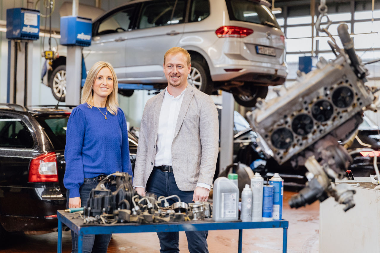 Lisa Woytinnek und Berater Florian Abschütz befinden sich in der Kfz‑Werkstatt des „Autohaus Kehm“. Im Hintergrund sind Fahrzeuge auf Hebebühnen sowie verschiedene Motor‑ und Ersatzteile im Servicebereich sichtbar, die den Werkstatt‑ und Wartungsbetrieb darstellen