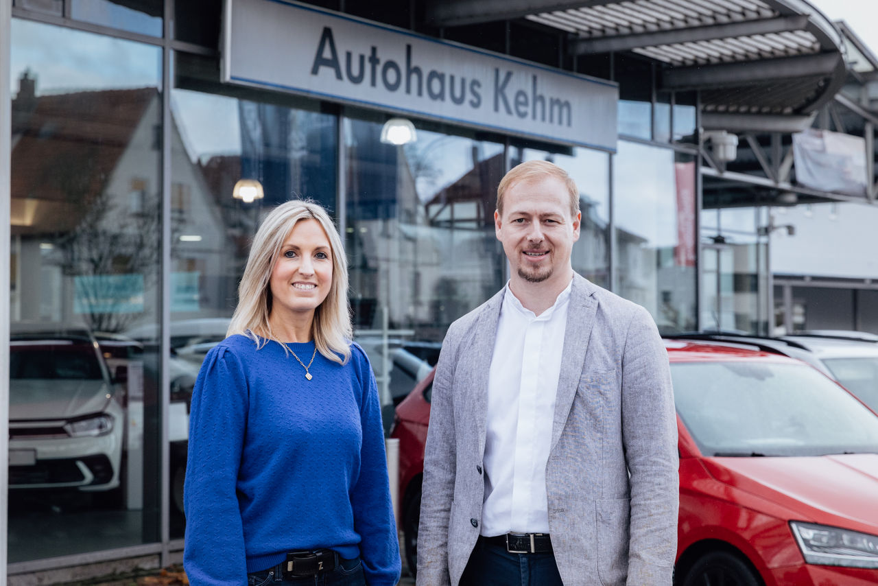 Lisa Woytinnek und Berater Florian Abschütz stehen vor dem „Autohaus Kehm“. Im Vordergrund ist der Fahrzeugbestand im Eingangsbereich zu sehen, im Hintergrund das Verkaufsgebäude des Autohauses, das den Standort und die Ausstellungsfläche nach außen präsentiert