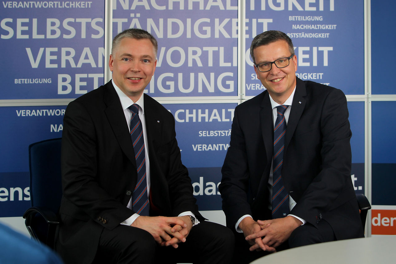 Zwei Männer in Anzügen, die auf Stühlen vor einer blauen Wand sitzen und sich unterhalten. 