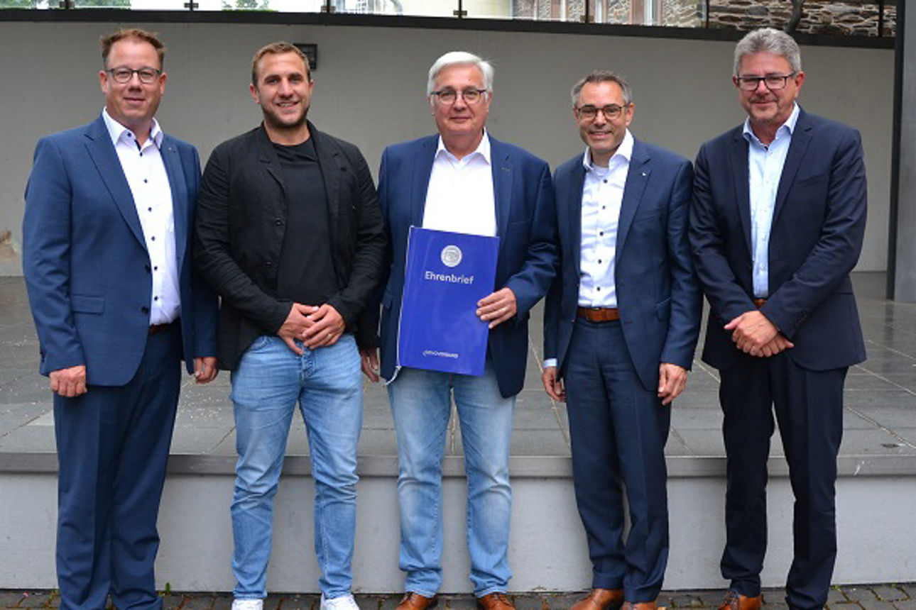 Fünf Männer stehen in einer Reihe, alle in Anzügen, vor einem grauen Wandhintergrund.