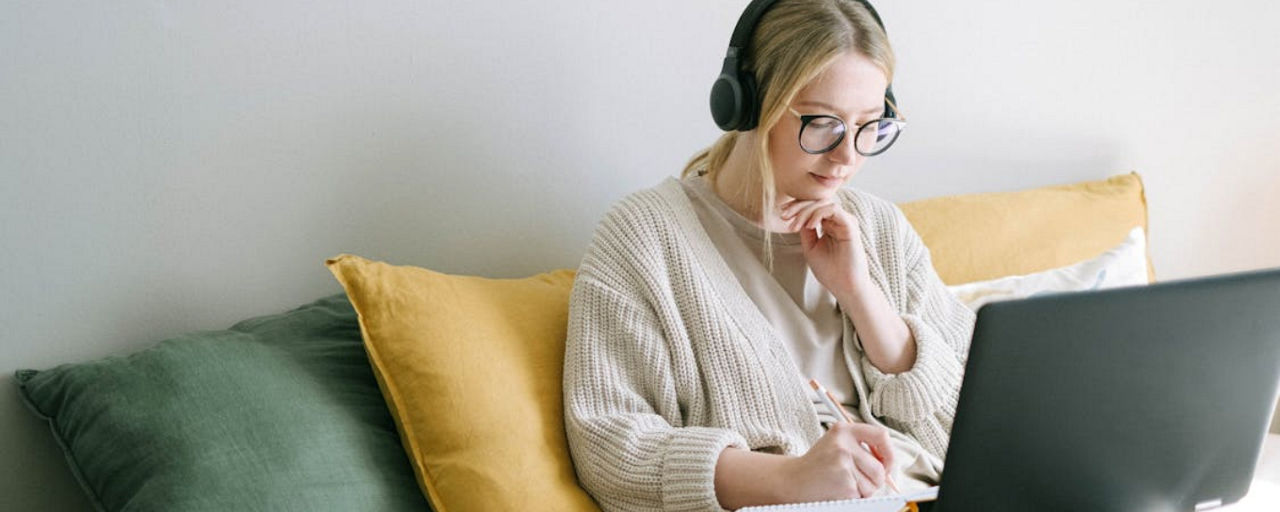 Eine Frau sitzt auf der Couch, hat ihren Laptop vor sich und Kopfhörer auf. 