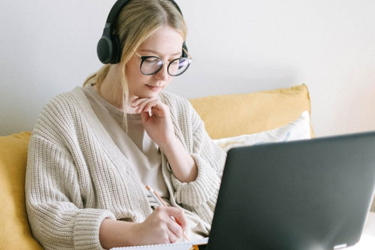 Eine Frau sitzt auf der Couch, hat ihren Laptop vor sich und Kopfhörer auf. 