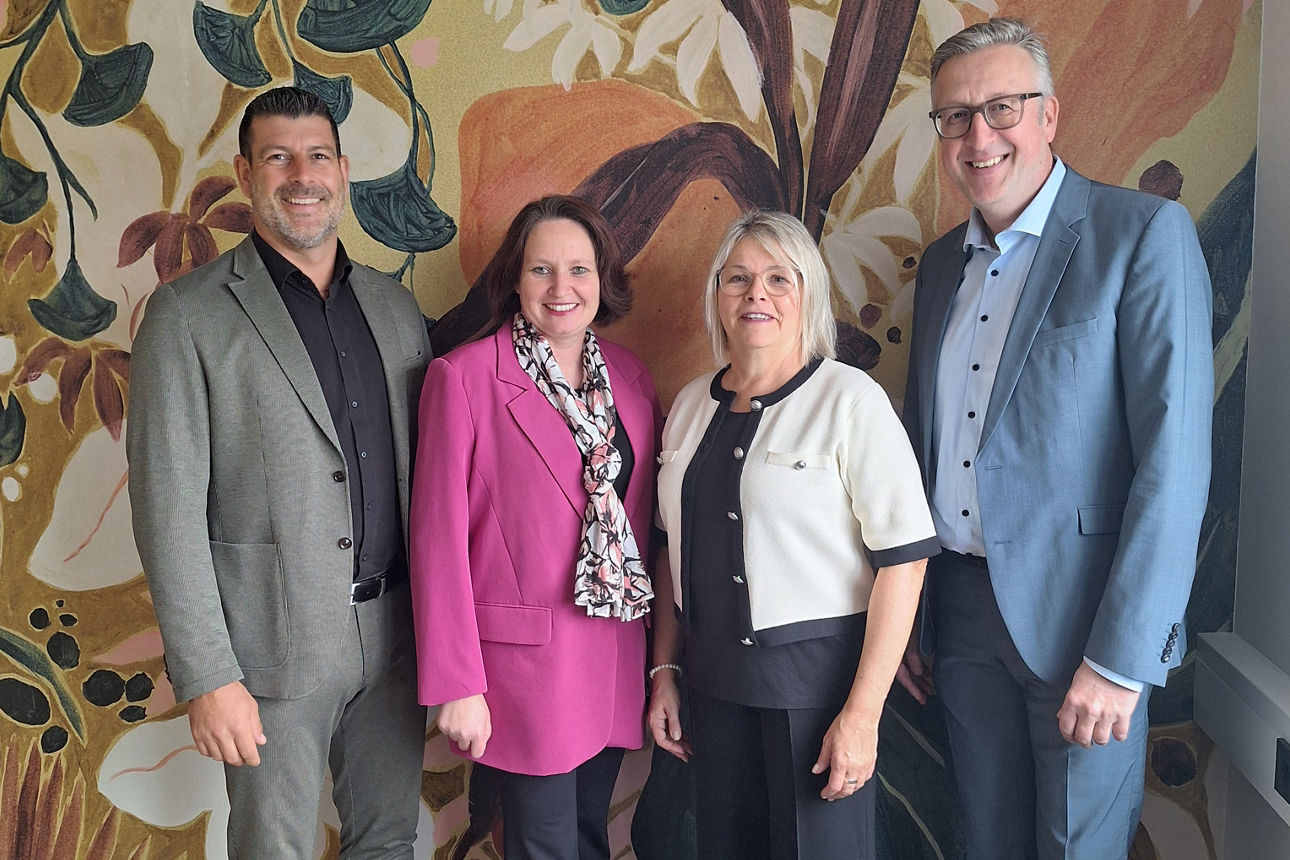 Auf dem Bild stehen zwei Männer und zwei Frauen vor einer floralen Tapete. Die Frauen stehen in der Mitte. Alle Personen lächeln in die Kamera.