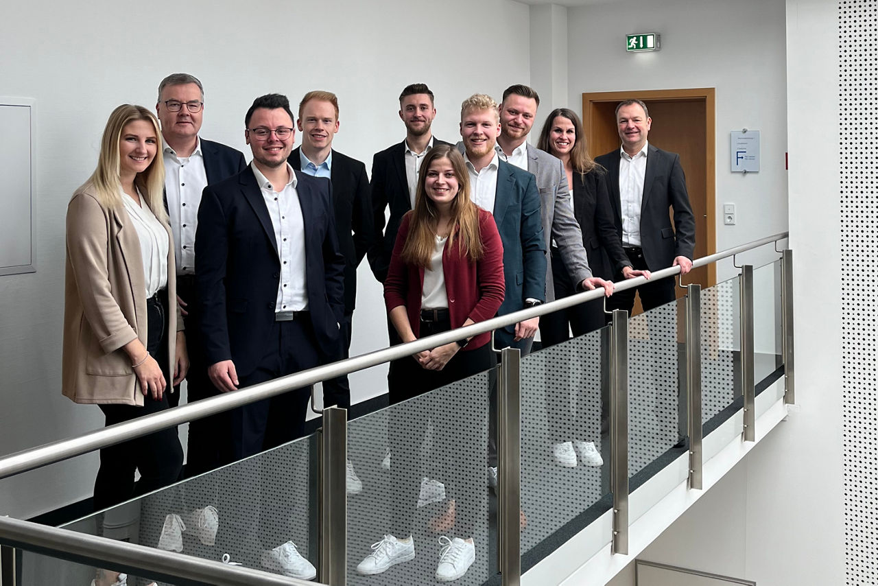 Gruppe von Absolventen eines berufsbegleitenden Studiums steht in einem modernen Gebäude an einem Geländer im Flur