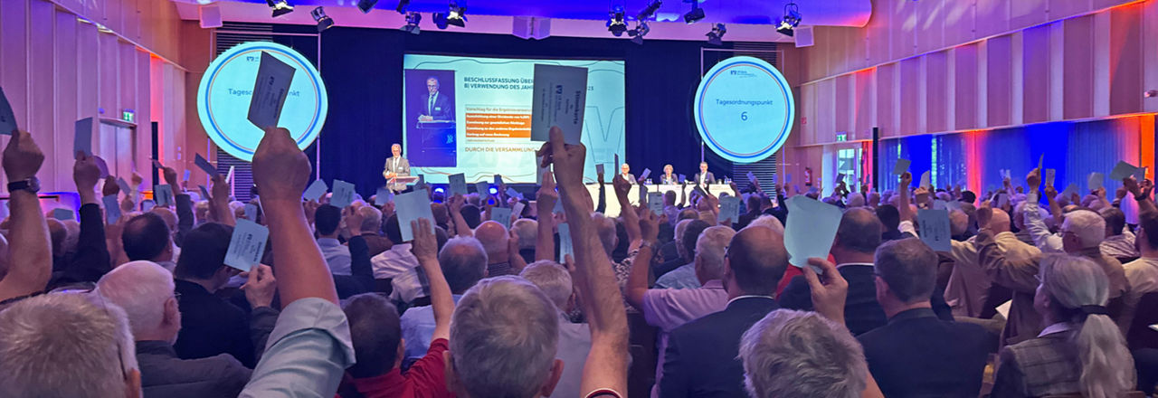 Das Bild zeigt die Vertreterversammlung der VR Bank. Anwesende stimmen ab, während eine Präsentation im Hintergrund sichtbar sind, die die Versammlung begleiten.