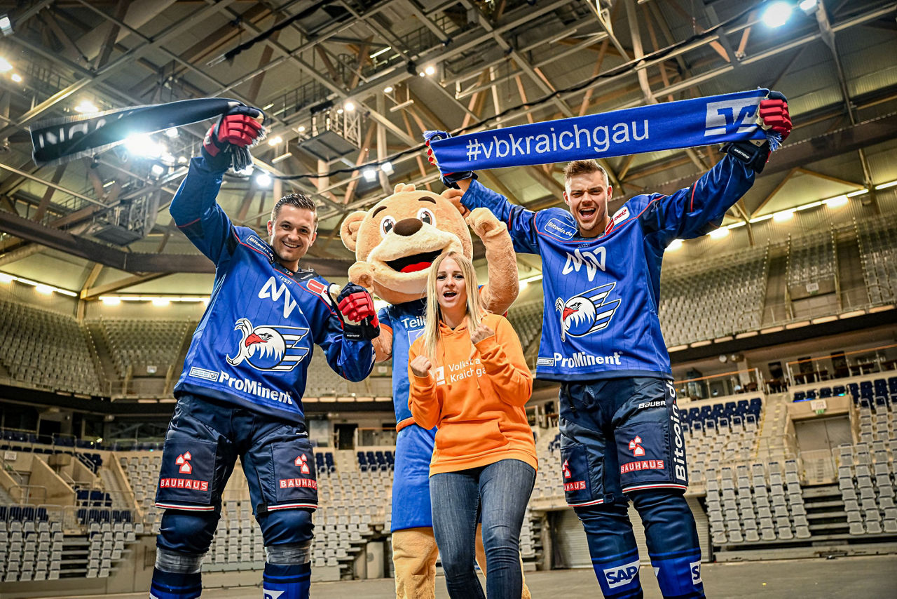 Zwei Spieler der Adler Mannheim, eine junge Frau im orangenen Pullover und unser TeilhaBÄR jubeln.