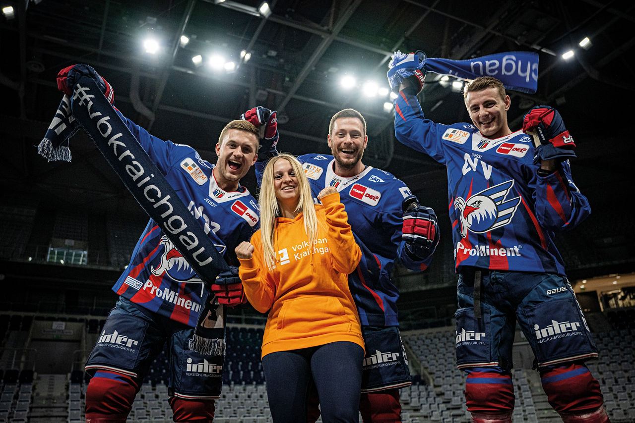 Drei Adler Mannheim Spieler und eine Volksbank Mitarbeiterin halten jubelnd einen Fanschal hoch.
