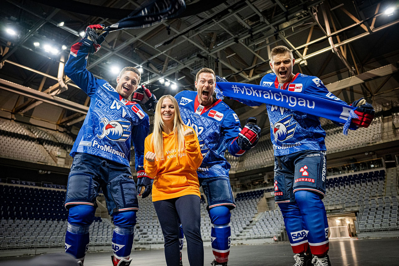 Drei Adler Mannheim Spieler und eine Volksbank Mitarbeiterin halten jubelnd einen Fanschal hoch.