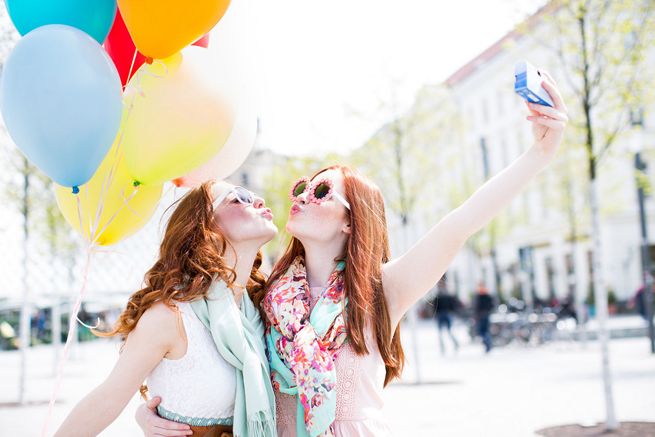 Zwei junge Frauen machen gemeinsam ein Selfie mit Kussmund. Im Hintergrund sind viele bunte Ballons