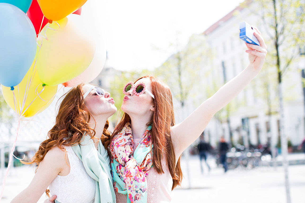 Zwei junge Frauen machen gemeinsam ein Selfie mit Kussmund. Im Hintergrund sind viele bunte Ballons