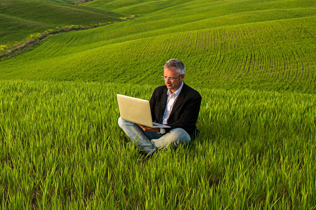 Mann sitzt inmitten eines grünen Feldes mit aufgeklappten Laptop
