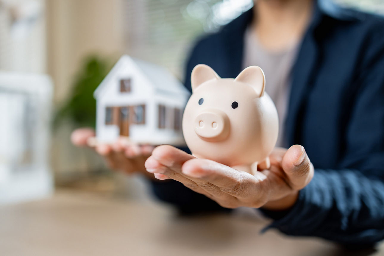 Eine Person hält ein Sparschwein in die Kamera und im Hintergrund hält die Person ein Modellhaus in der anderen Hand