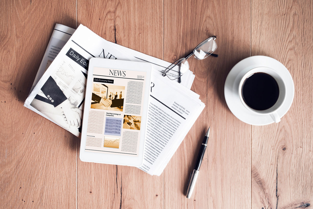 Auf einem Holztisch liegen Zeitungen, eine Brille, ein Tablet mit geöffneter Nachrichtenseite, ein Stift und eine Tasse Kaffee. Die Szene symbolisiert aktuelle Nachrichten oder Presseinformationen.