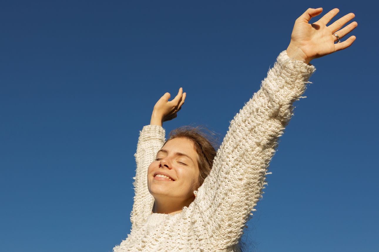 Eine glückliche junge Europäerin in ihren Zwanzigern genießt einen Moment in der Sonne unter wolkenlosem Himmel. Ihre Arme sind nach oben gestreckt.