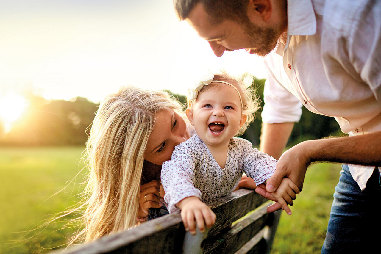 Ein glückliches Paar mit ihrer kleinen Tochter, die sich lachend an ihrem Vater festhält. Im Hintergrund geht die strahlende Sonne unter.