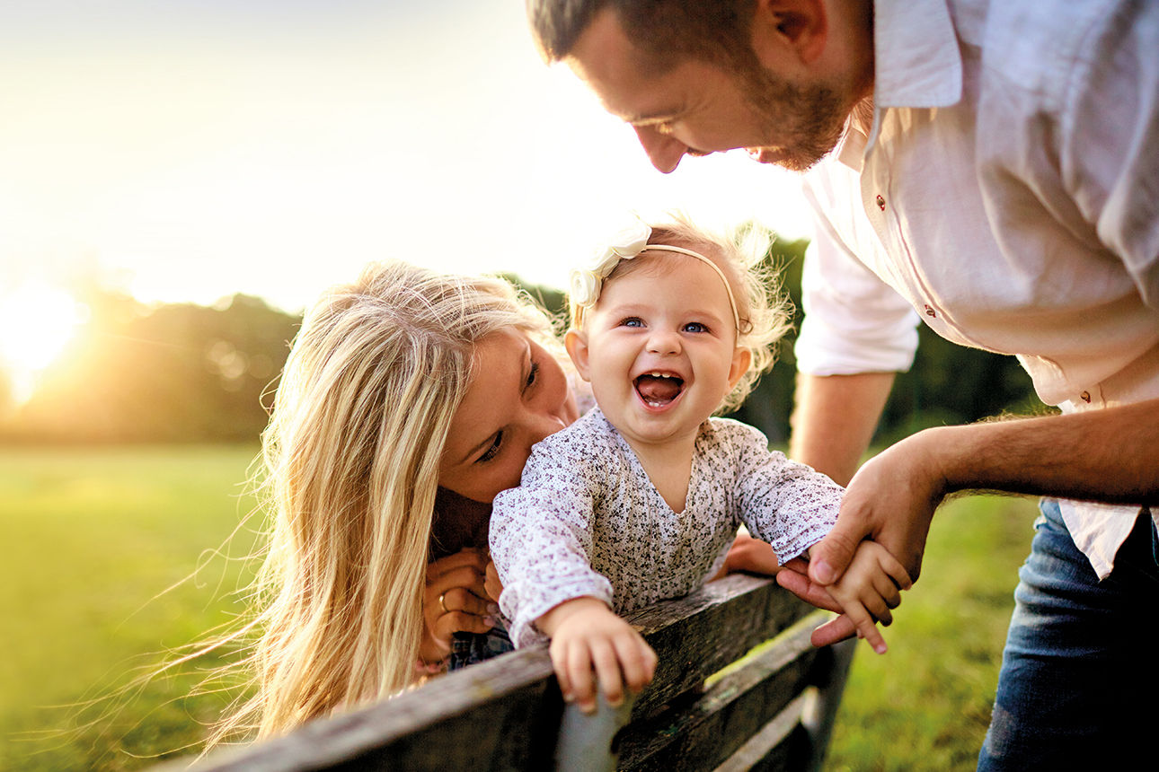 Ein glückliches Paar mit ihrer kleinen Tochter, die sich lachend an ihrem Vater festhält. Im Hintergrund geht die strahlende Sonne unter.
