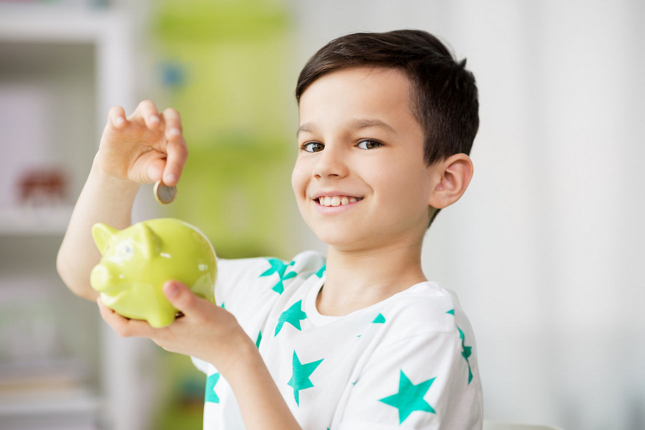 money, finances, childhood and people concept - smiling little boy putting coin into piggy bank at home
