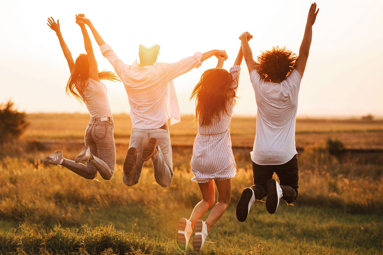 Two young guys and two girls are holding their hand and jumping in the field on a summer day. Back view.