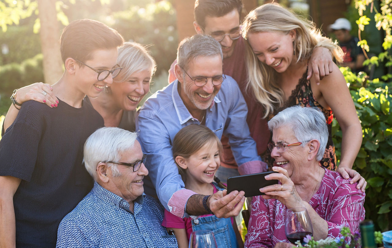 Im Sommer vergnügt sich eine Familie mit drei Generationen an einem Tisch im Garten und isst gemeinsam. Alle Gäste schauen sich ein Video auf dem Handy an.