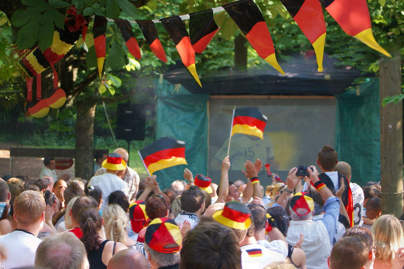 Menschenmenge im Freien mit Deutschlandfahnen, Deutschlandmützen und Deutschlandwimpeln bei einer Veranstaltung unter Bäumen.