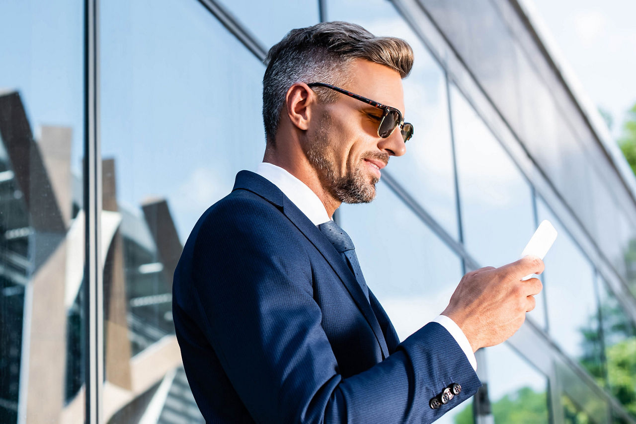 Mann im Anzug mit Sonnenbrille schaut lächelnd auf sein Handy