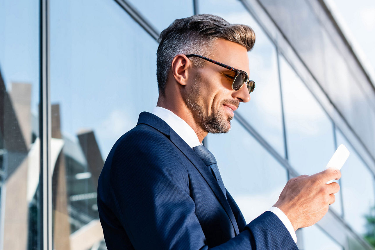 Mann im Anzug mit Sonnenbrille schaut lächelnd auf sein Handy
