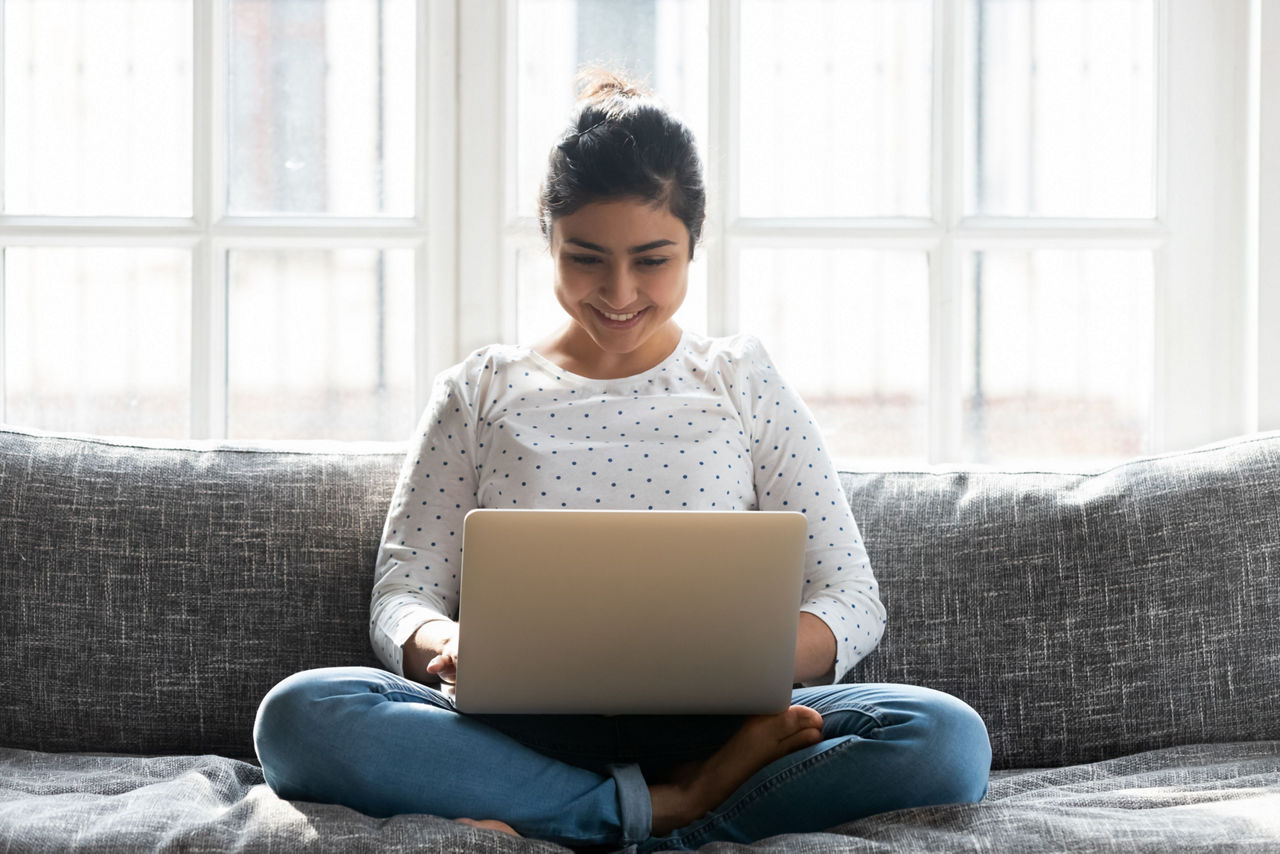Lächelnde junge Frau sitzt mit ihrem Laptop im Schneidersitz auf dem Sofa.