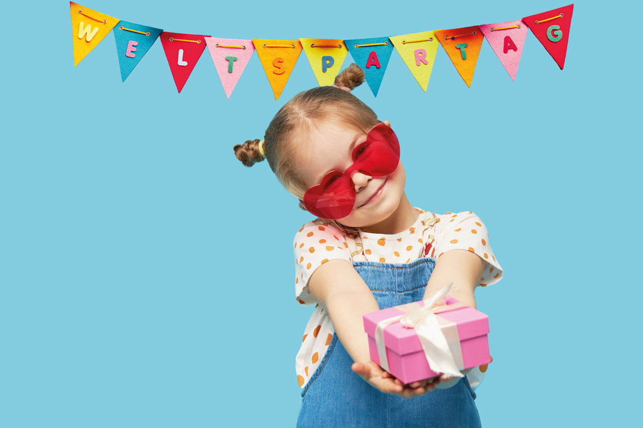 Ein Mädchen mit einem Geschenk in der Hand steht vor einer Wimpelkette mit der Aufschrift "Weltspartag"