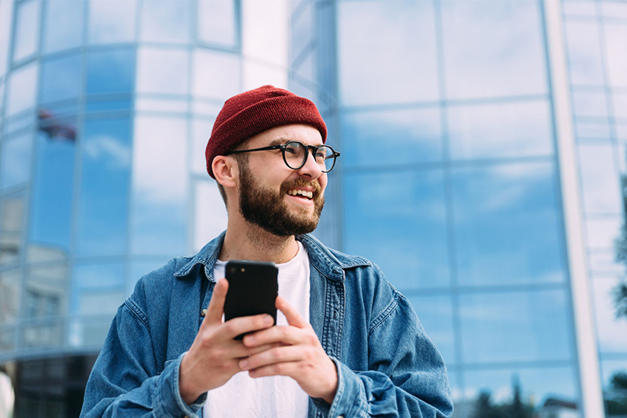 Junger Mann steht mit einem Smartphone in der Hand vor einem Gebäude mit Glas
