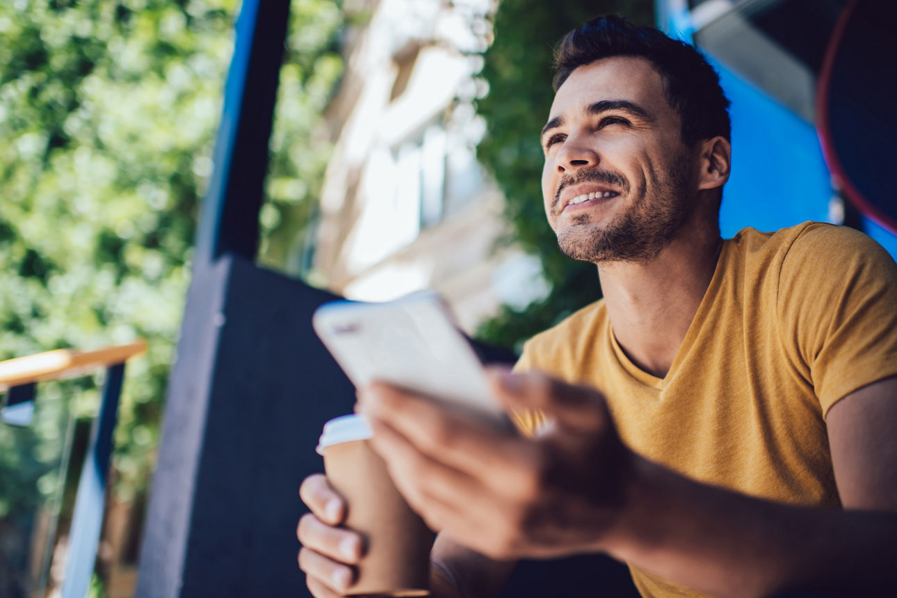 Ein junger Mann sitzt draußen mit einem Kaffee und liest auf seinem Handy.