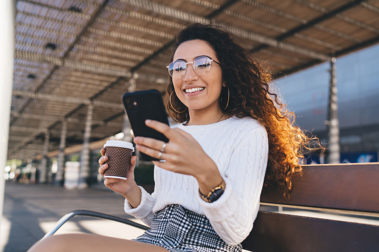 Frau sitzt mit Kaffebecher in der Hand auf einer Bank und schaut lächelnd auf ihr Smartphone